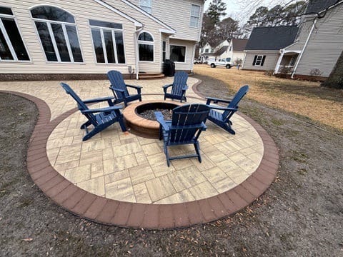 Curved paver fire pit patio with blue Adirondack chairs behind a home