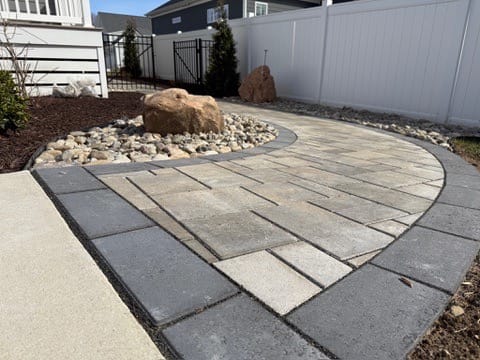 Curved paver walkway with river rock and boulder beside a white fence