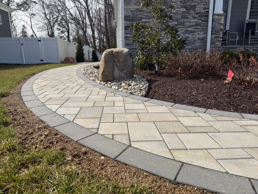 Light paver walkway with dark border beside a boulder and mulch bed