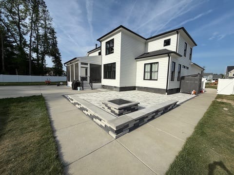 Raised paver patio with square fire pit beside a modern white home