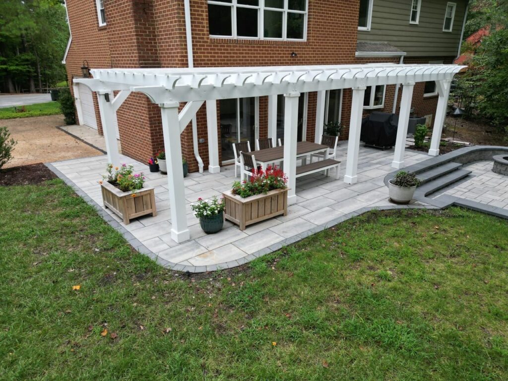 White pergola over a paver patio with planters beside a brick home